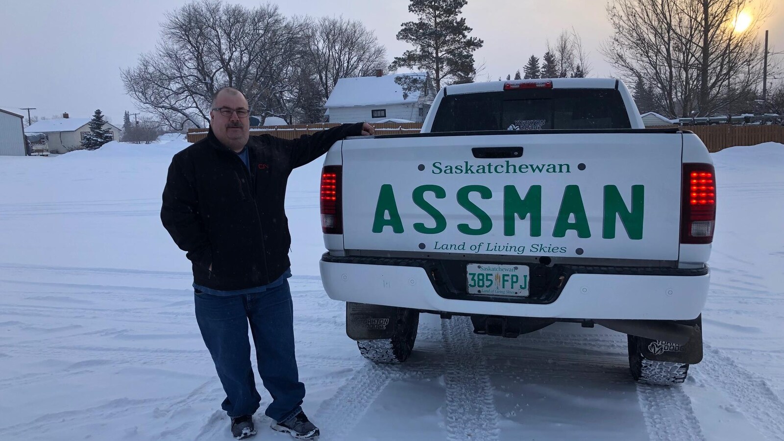 Un homme pose avec sa camionnette blanche où on peut voir un autocollant qui imite la plaque d'immatriculation de la Saskatchewan et où il est inscrit Assman.