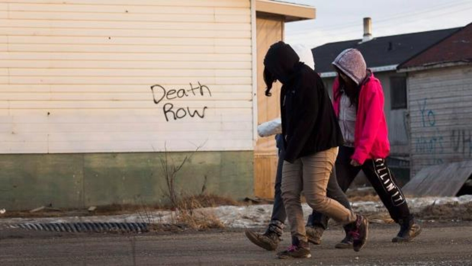 Trois jeunes marchent dans une rue de la communauté crie d'Attawapiskat. Derrière eux, l'inscription « Death Row » apparaît sur le mur d'une maison.