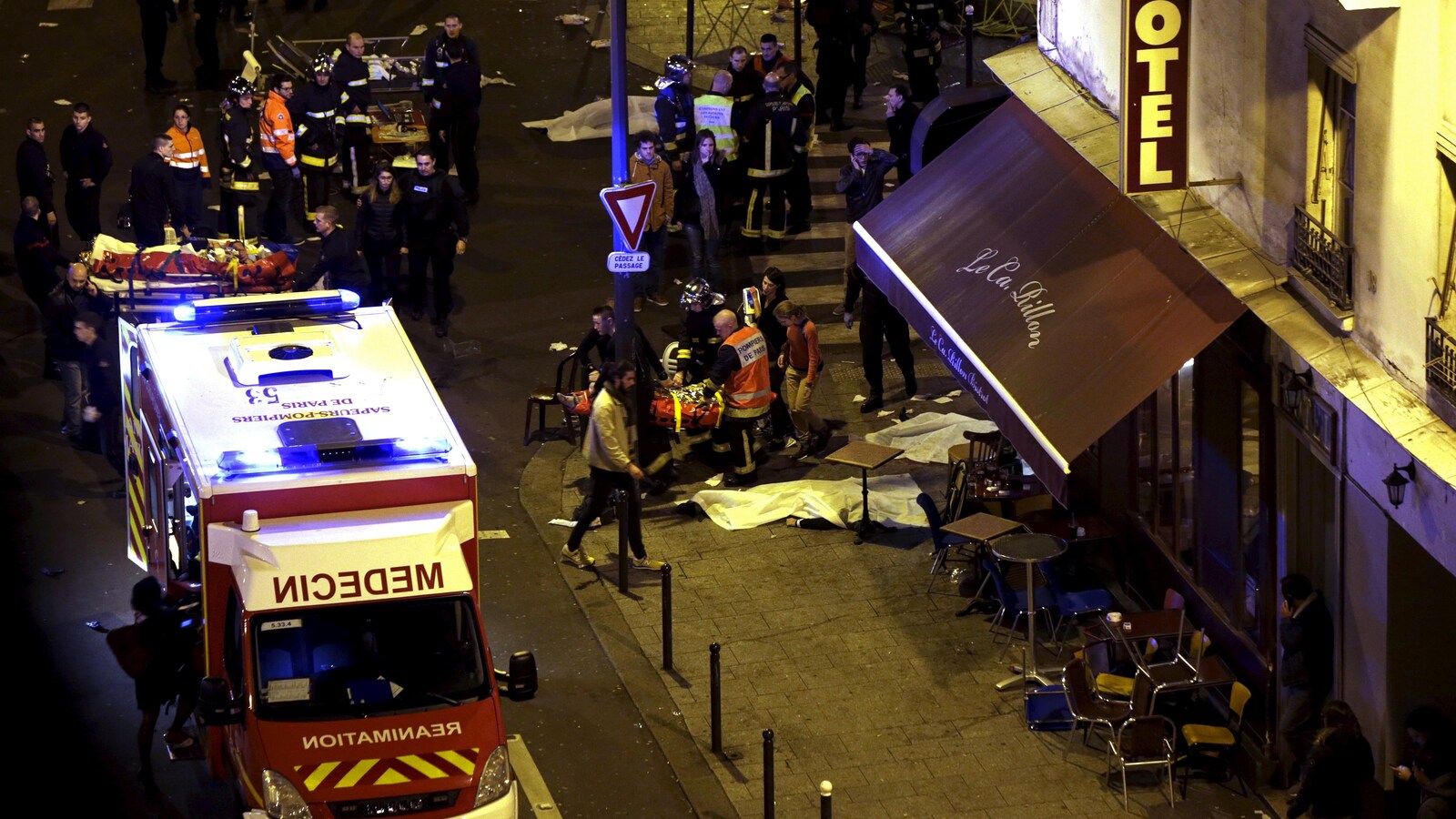 Vue d'ensemble d'un coin de rue où des policiers et des secouristes s'affairent autour d'un camion de pompiers, quelques bâches cachant des corps.
