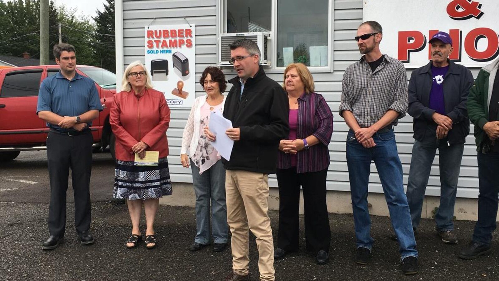 Kris Austin en conférence de presse entouré de quelques partisans devant une imprimerie.