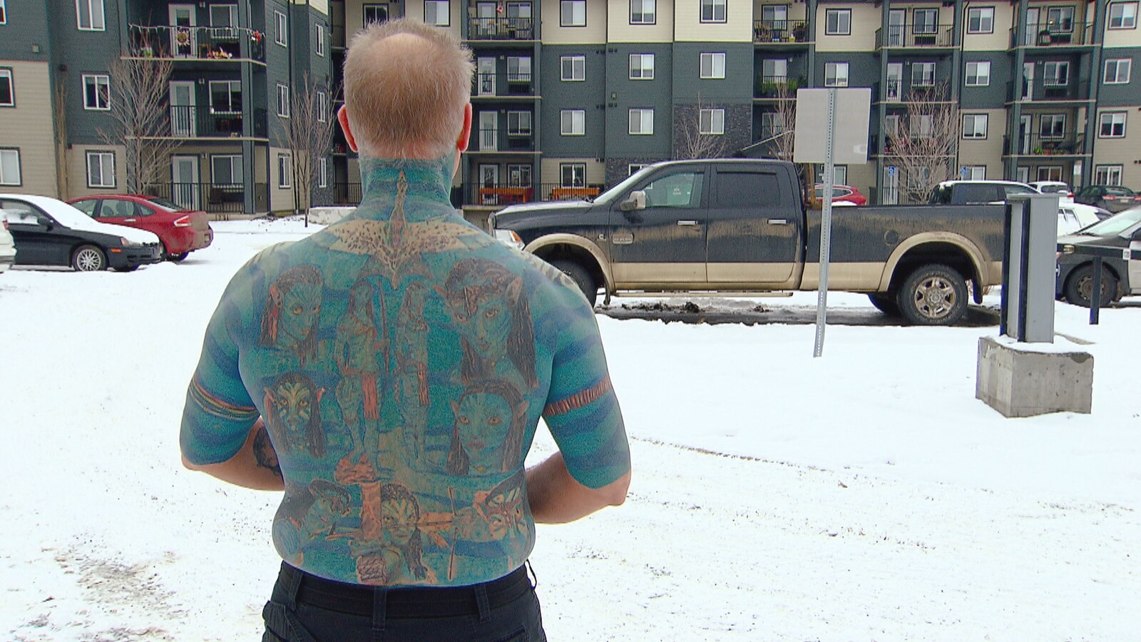 Un homme est debout de dos, à l'extérieur. Il est couvert de tatouages bleus de la nuque aux coudes et à la taille.