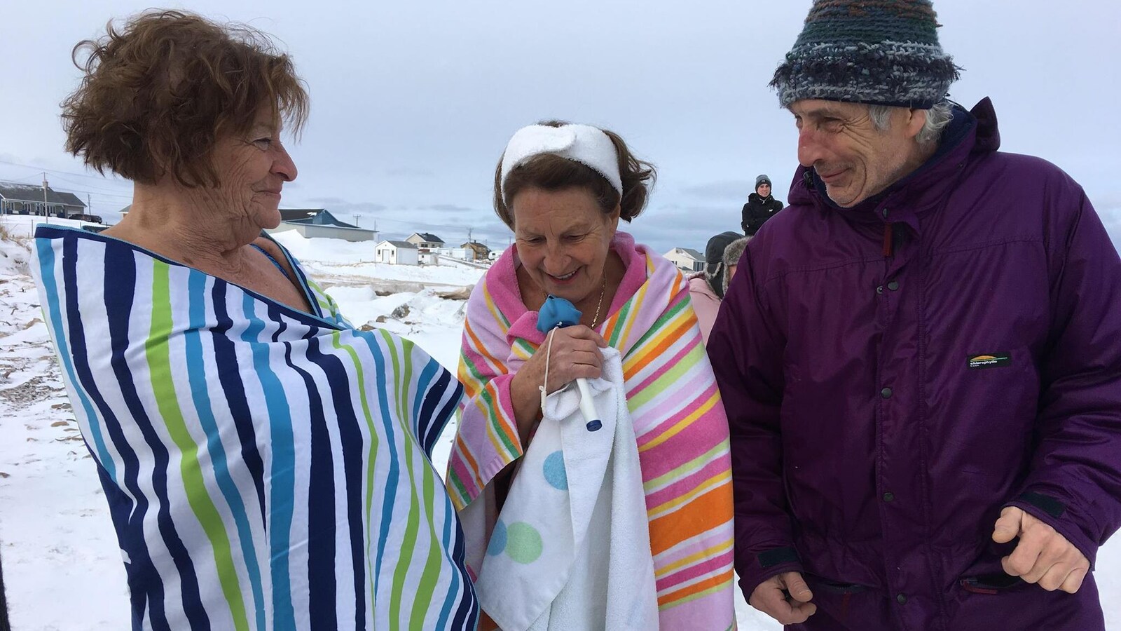 On voit deux femmes et un homme dehors, pendant l'hiver. Ils sont sur une plage recouverte de neige aux Îles-de-la-Madeleine. Les deux femmes sont enroulées dans des serviettes, car elles se sont jetées à l'eau dans le golfe du Saint-Laurent.