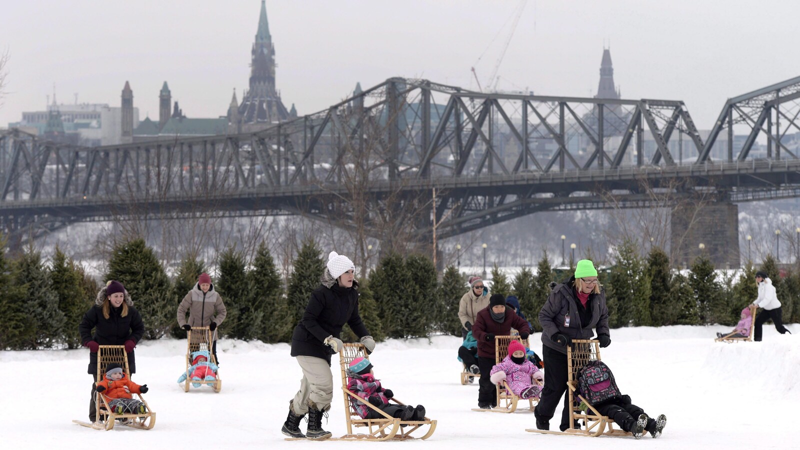 Au premier plan, on peut voir des parents en train de pousser leurs enfants dans des traîneaux sur la glace du canal Rideau, avec en arrière plan, le Parlement d'Ottawa.