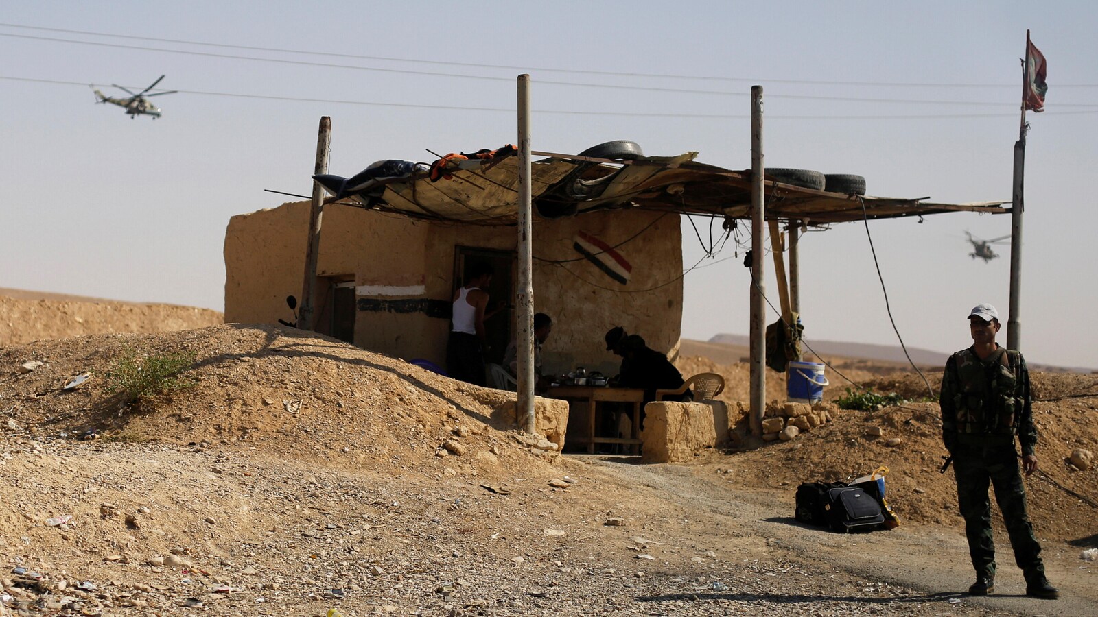 Un hélicoptère russe survole un bâtiment rudimentaire gardé par un homme en habit militaire à la base aérienne de Chaïrat, au sud-est de Homs, en Syrie.