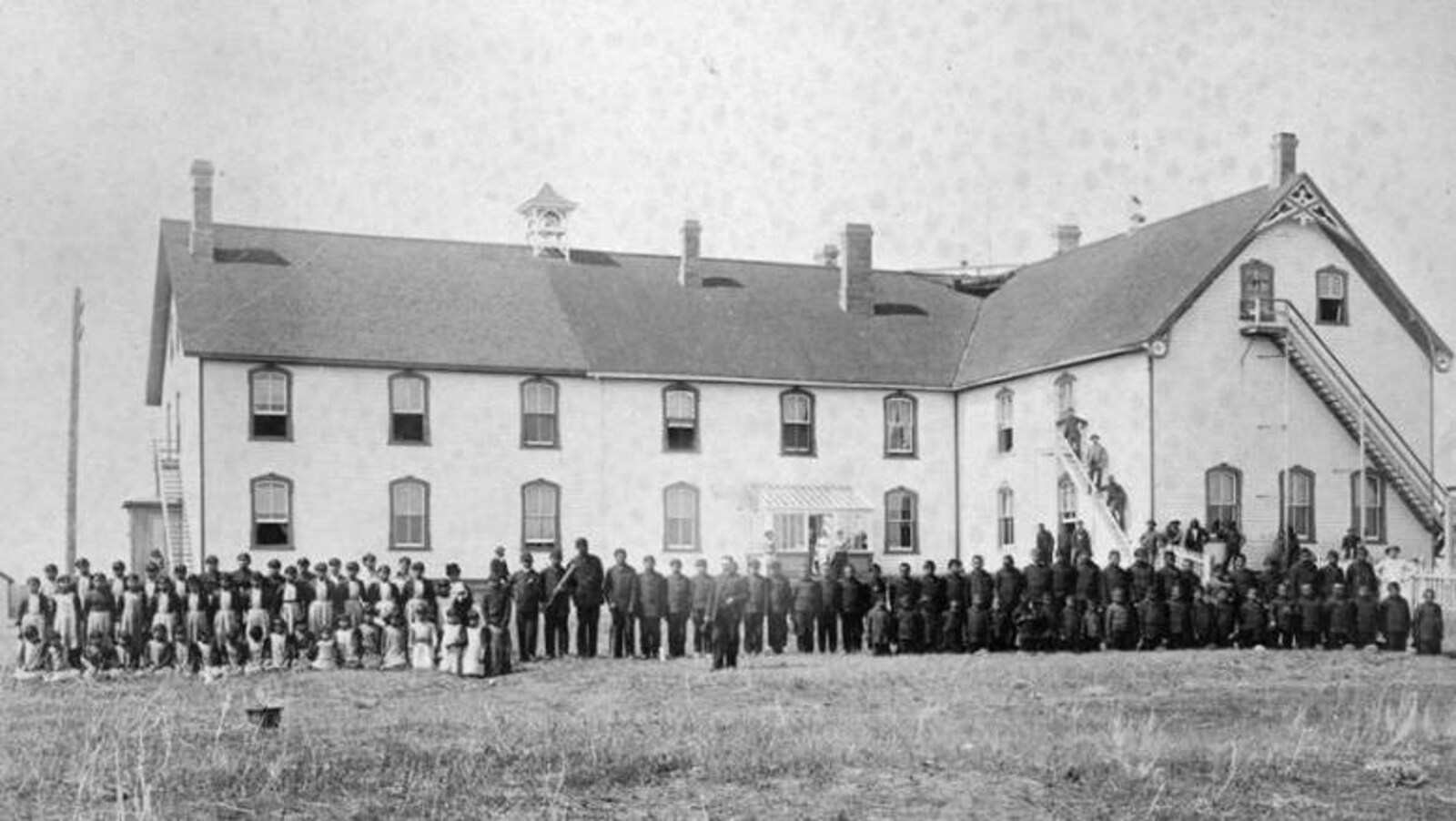 Des élèves debout devant le pensionnat autochtone Battleford Industrial School en 1895. Un édifice à deux étages.