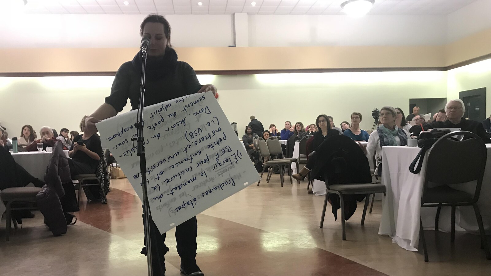 Dans une salle comblée, une femme derrière un microphone lit ce qui est écrit sur une immense feuille de papier.