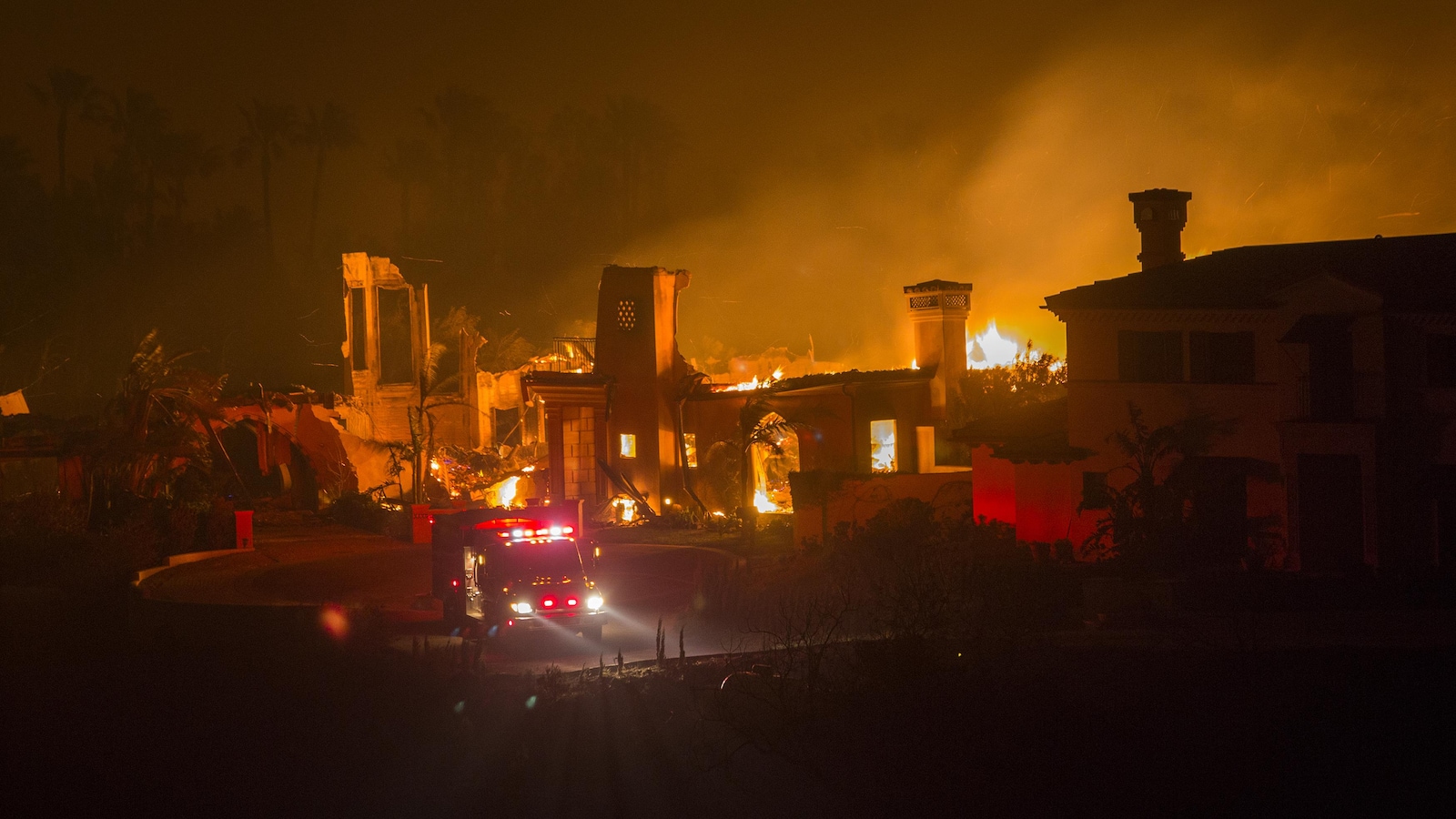 En pleine nuit, un feu de forêt brûle une maison devant laquelle se trouve un camion de pompiers.