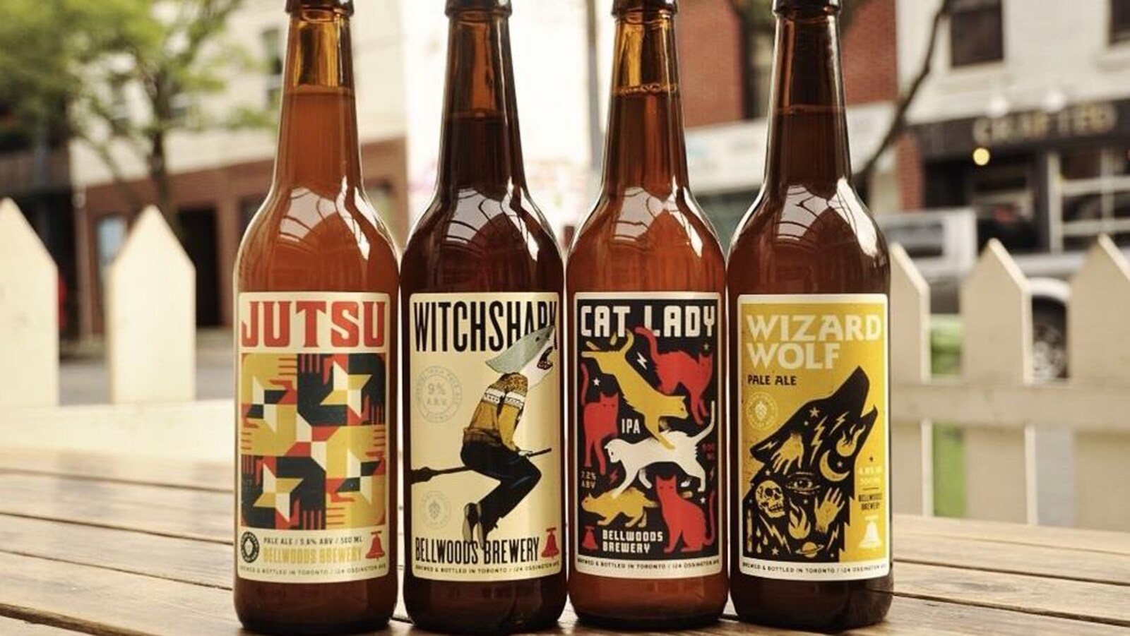 Quatre bouteilles de différentes bières vendues par la brasserie Bellwoods sont alignées sur une table en bois, dans une rue.