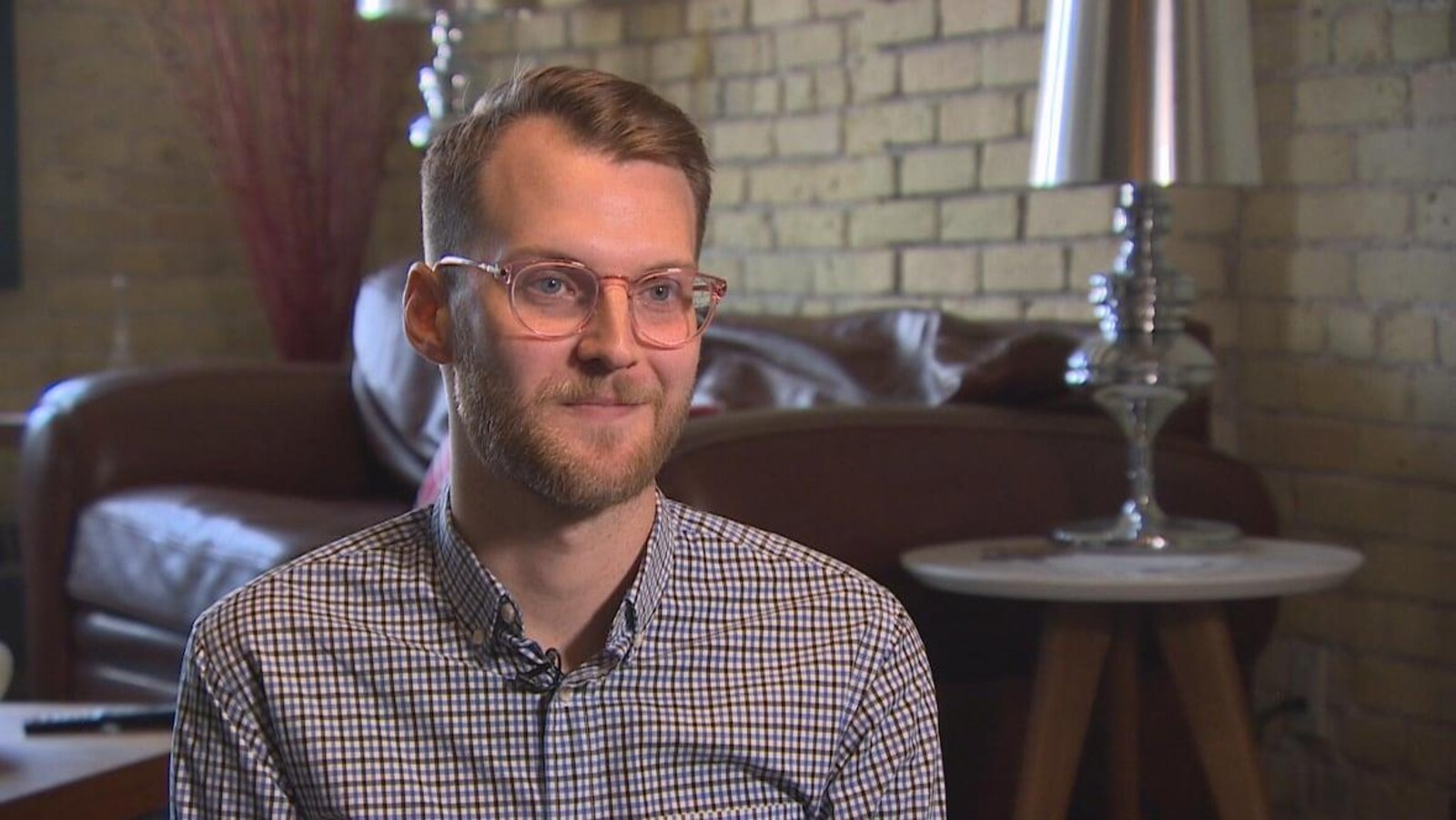 Un jeune homme blond à lunettes, dans un salon.