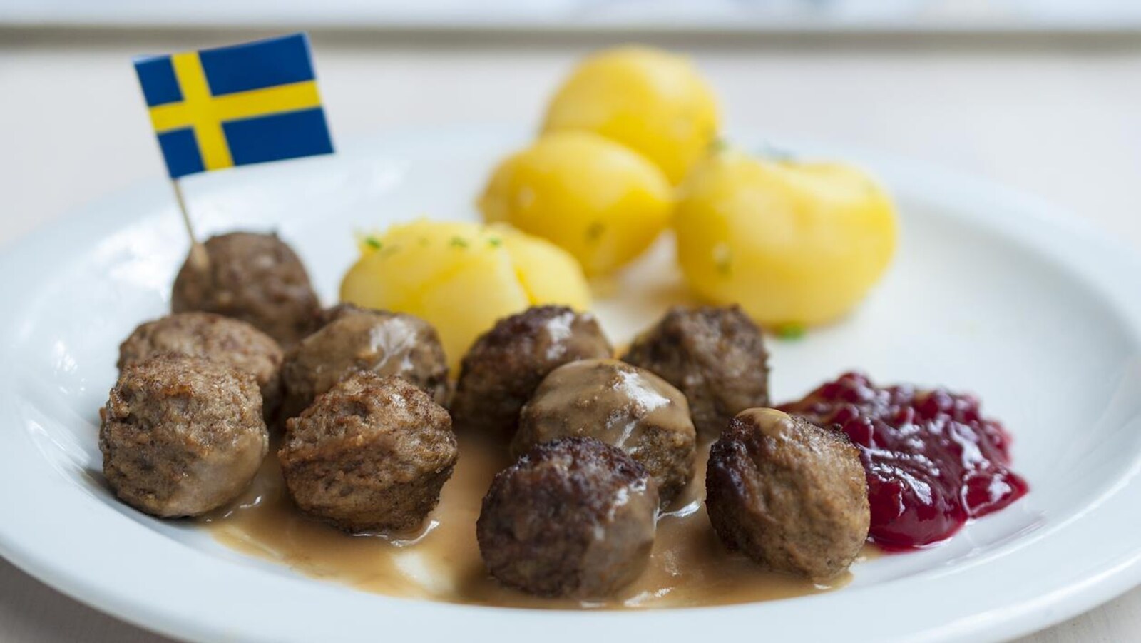 Des boulettes de viande servies dans une assiette accompagnées de pommes de terre, de sauce brune et de sauce aux canneberges, décorées d'un petit drapeau de la Suède.