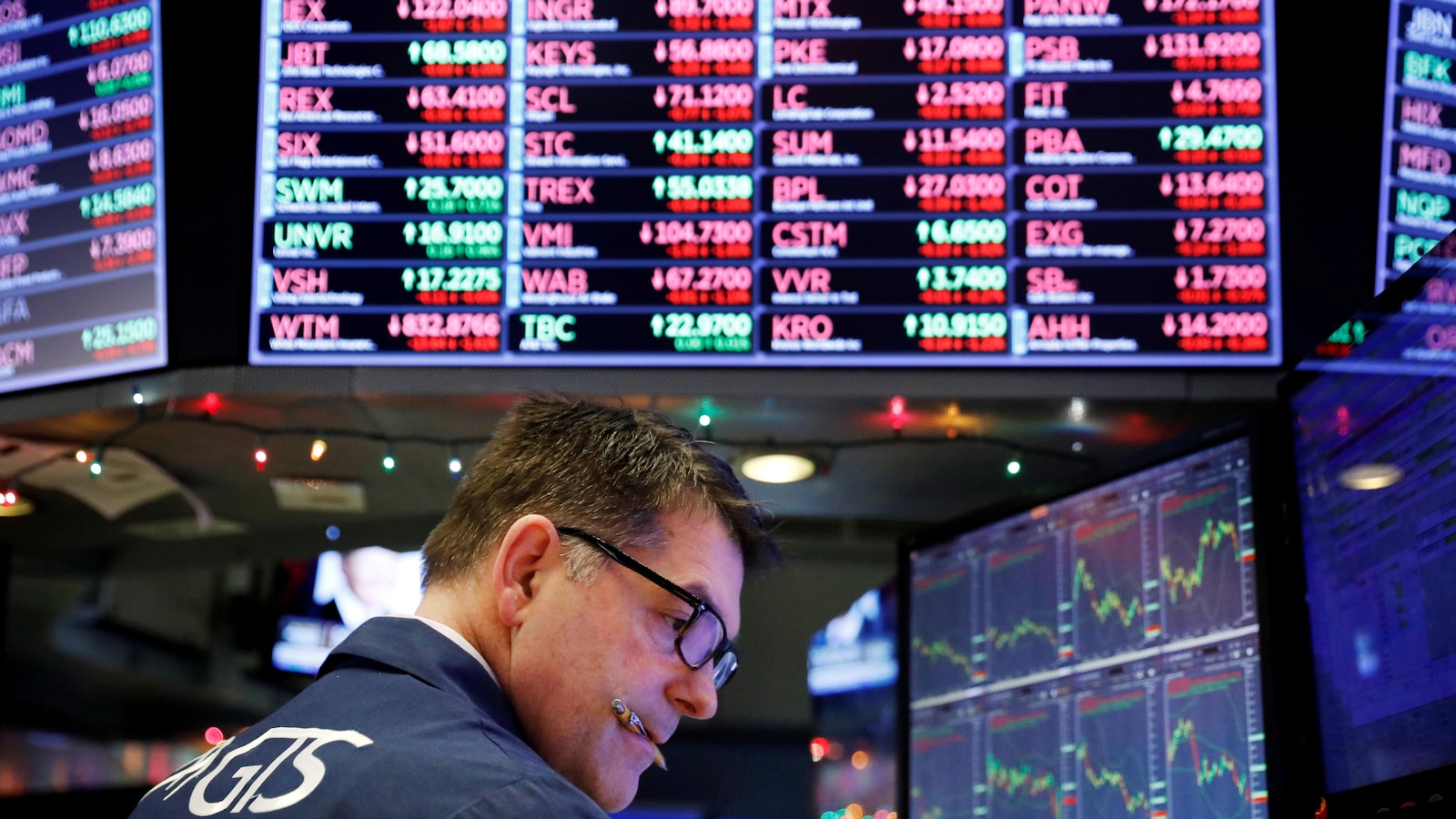 Un homme a un stylo dans la bouche alors qu'il consulte le cours de la bourse sous un énorme écran sur lequel sont affichées diverses actions de compagnies.