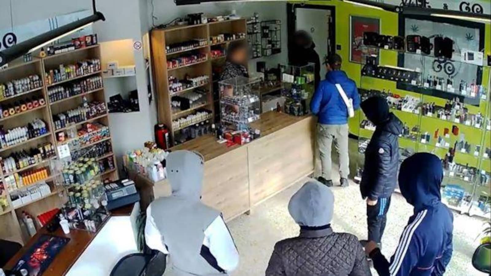 Dans un magasin, on distingue des cigarettes électroniques. Cinq individus, capuche sur la tête et le dos tourné, font face à un vendeur.