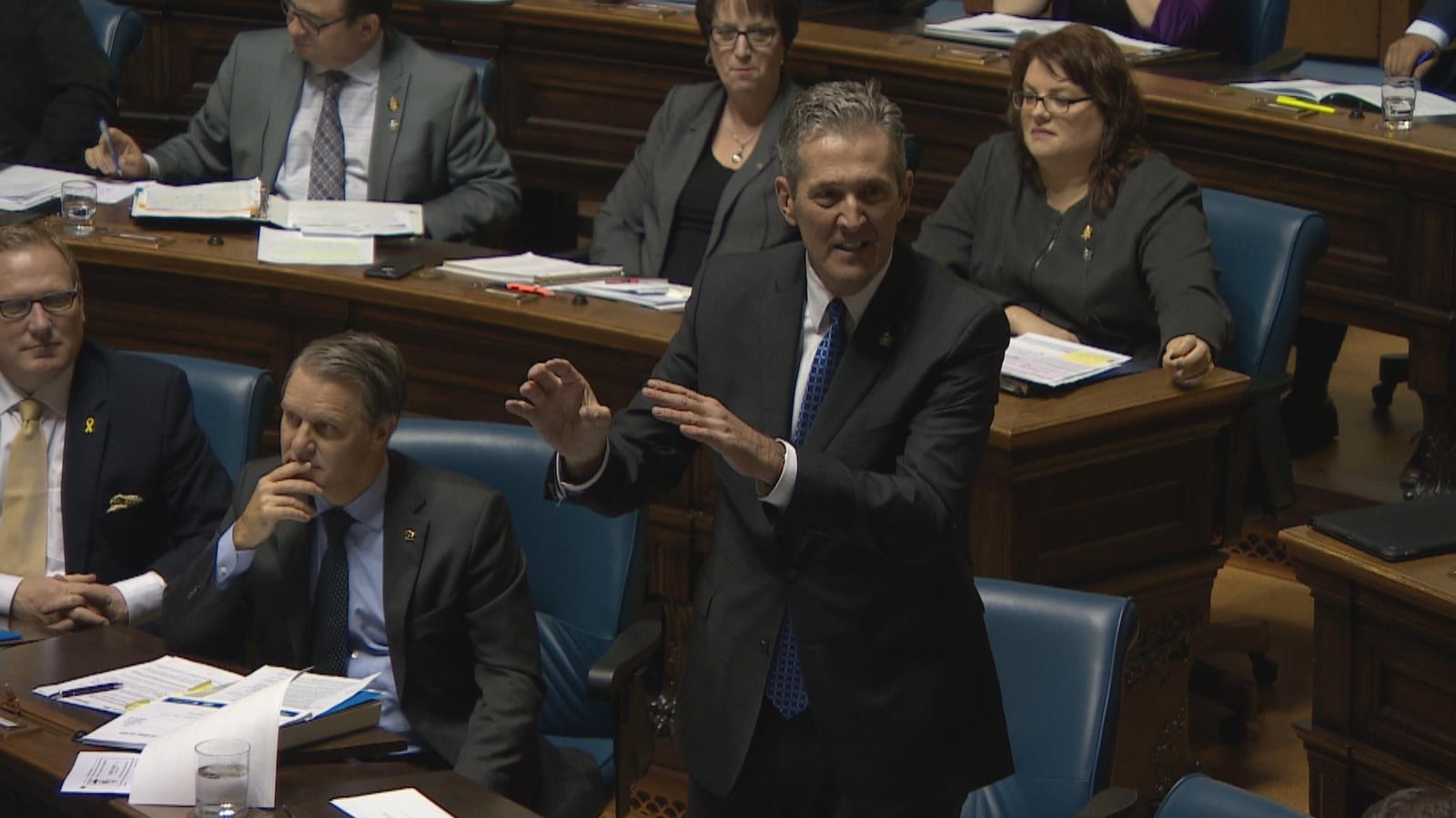 Pallister, debout en chambre, agite les mains en répondant à une question. À côté de lui, des ministres du gouvernement sont assis et écoutent.
