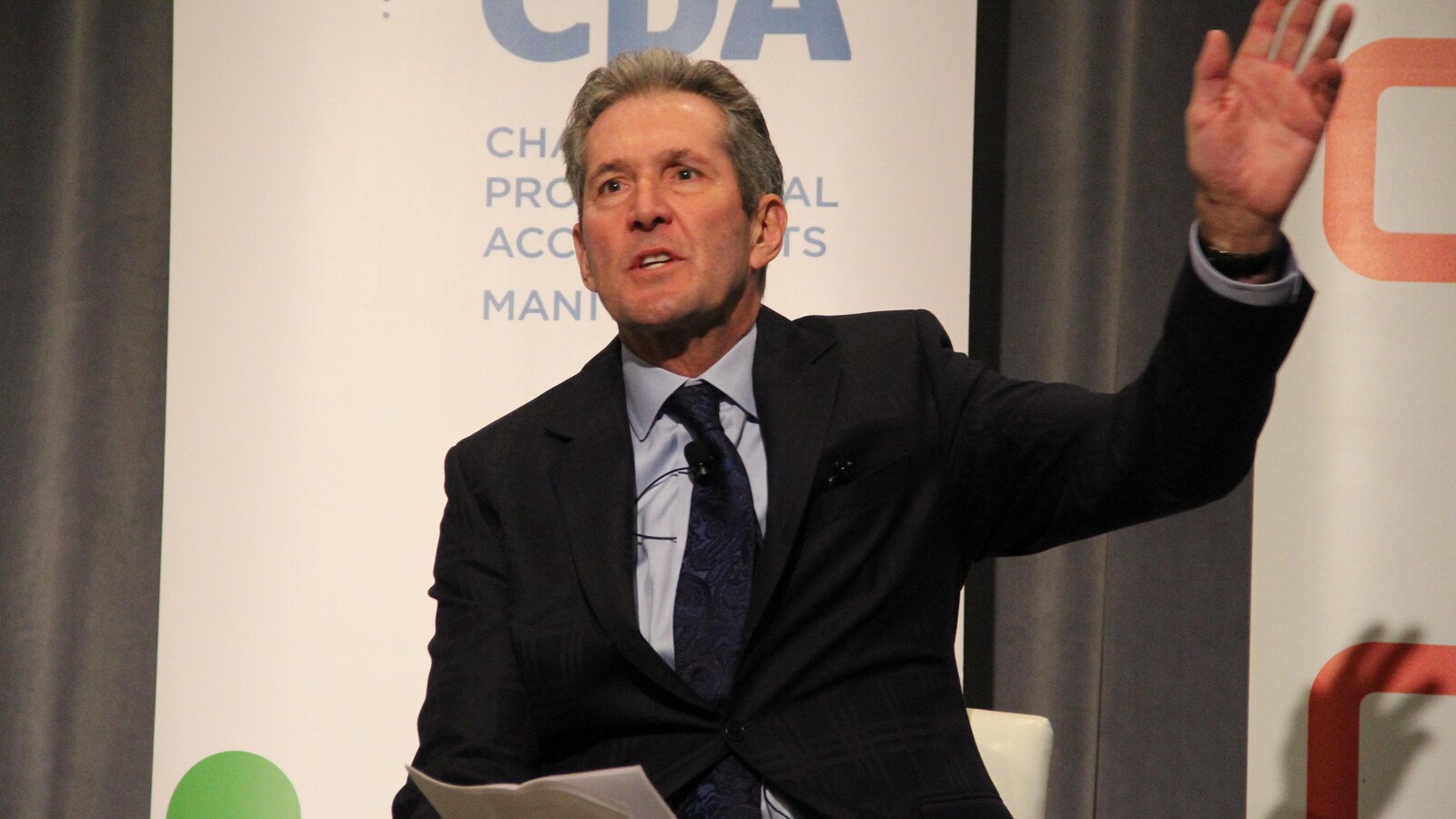 Le premier ministre du Manitoba, Brian Pallister, est photographié alors qu'il parle. Il est assis, tient des feuilles de papier dans sa main droite et lève la main gauche tout en s'exprimant.