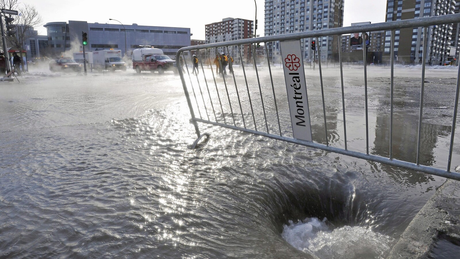 Vue panoramique d'une intersection inondée, une barrière métallique ornée du logo de la Ville de Montréal au premier plan.