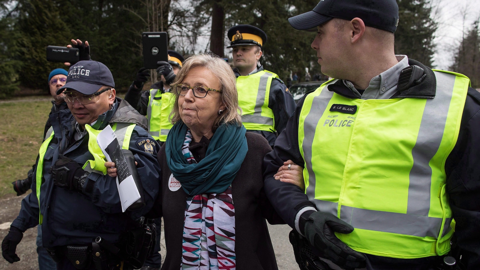 Elizabeth May est encadrée par deux policiers et suivie par deux photographes et d'autres policiers.
