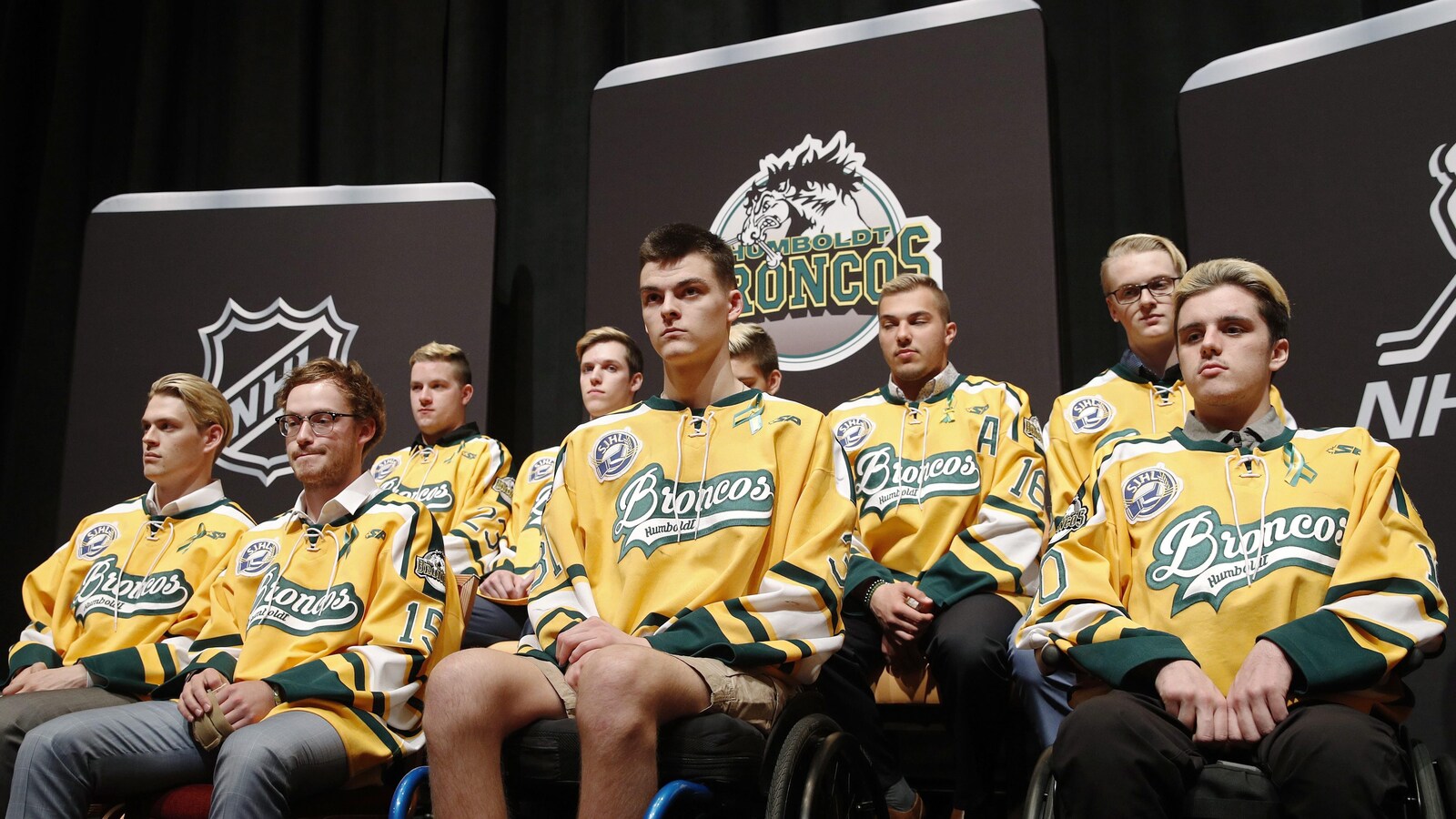 Les joueurs survivants se tiennent devant des affiches de la LNH et des Broncos de Humboldt. Ils portent tous des chandails de leur équipe de hockey.