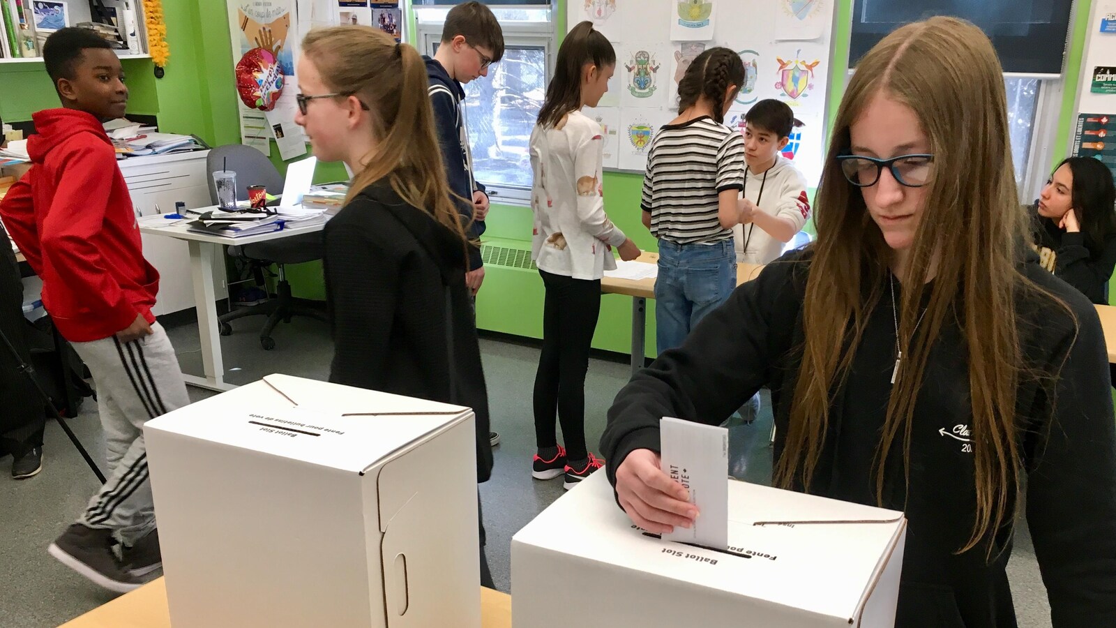 Une jeune fille aux cheveux longs et à lunettes dépose un papier dans la fente d'une boîte en carton posée sur une table. Une autre boite en carton est posée à côté. Derrière, des jeunes sont en ligne et semblent attendre leur tour.