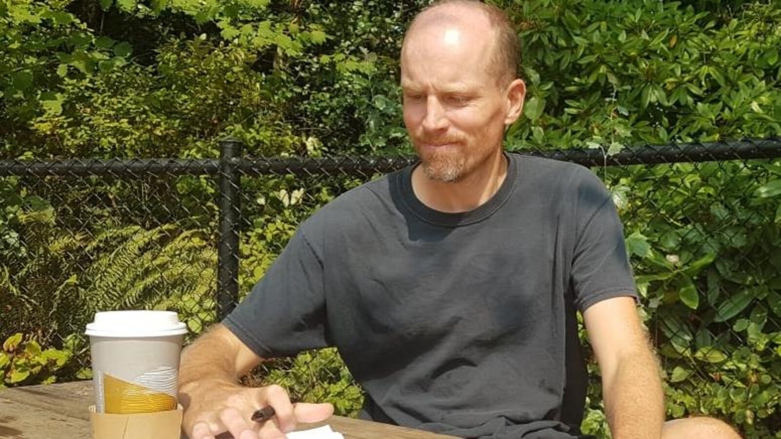 Un homme le crâne dégarni assis à une table de pique-nique avec un stylo à la main et une tasse de café posé sur son calepin
