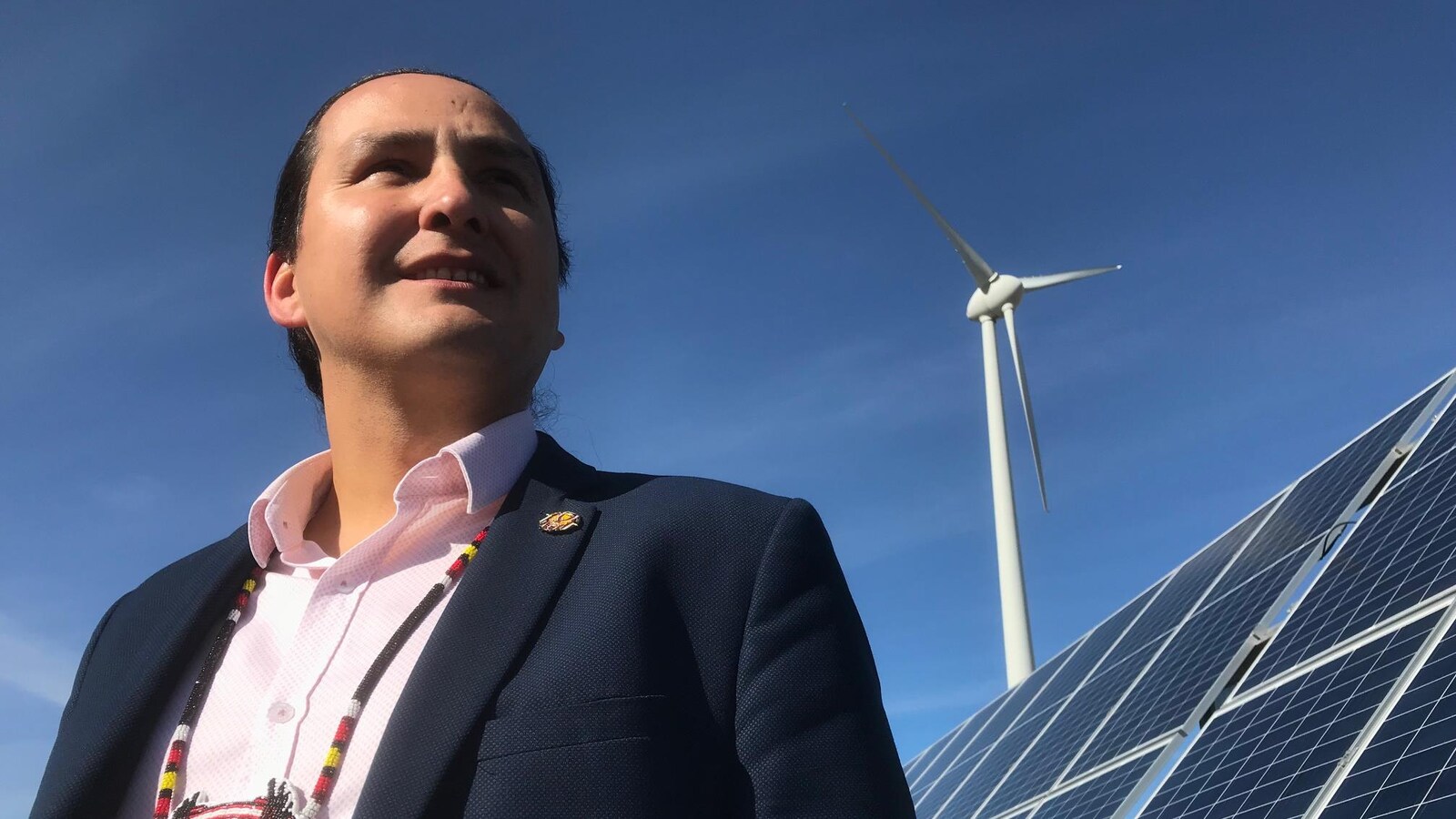 Un homme avec un collier traditionnel autochtone devant un panneau solaire et une éolienne