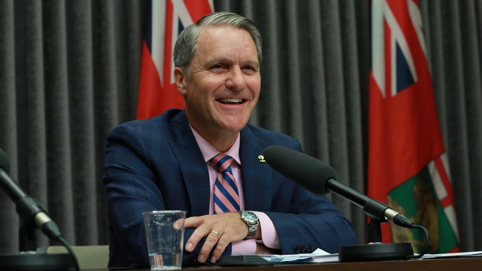 Un homme souriant à une table avec un micro, et des drapeaux de la province du Manitoba derrière lui.