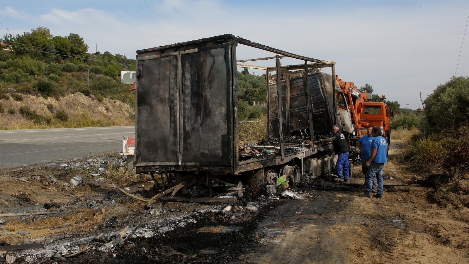 Trois personnes près d'un camion calciné, sur le bord d'une route en campagne.