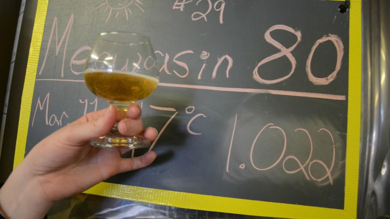 Une main de femme tient un verre de bière devant un tableau où il est écrit à la craie : Meewasin 80, Mar. 11, 17 degrés, 1.022.