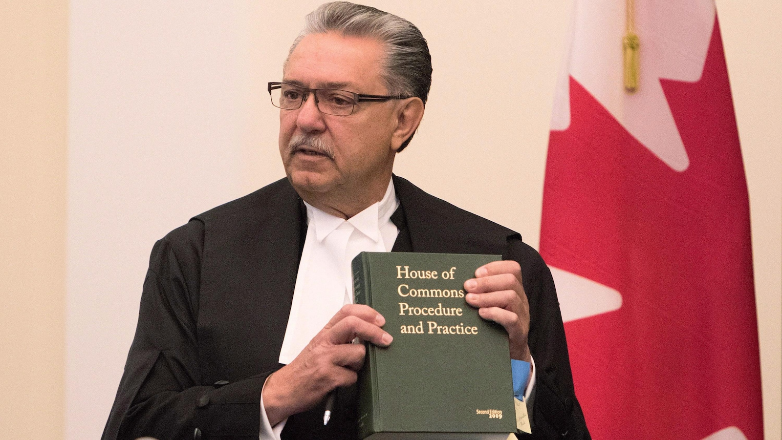 Gene Zwozdesky s'adresse aux députés, tenant en main le livre des Normes et procédures de la Chambre des communes, devant un drapeau du Canada.