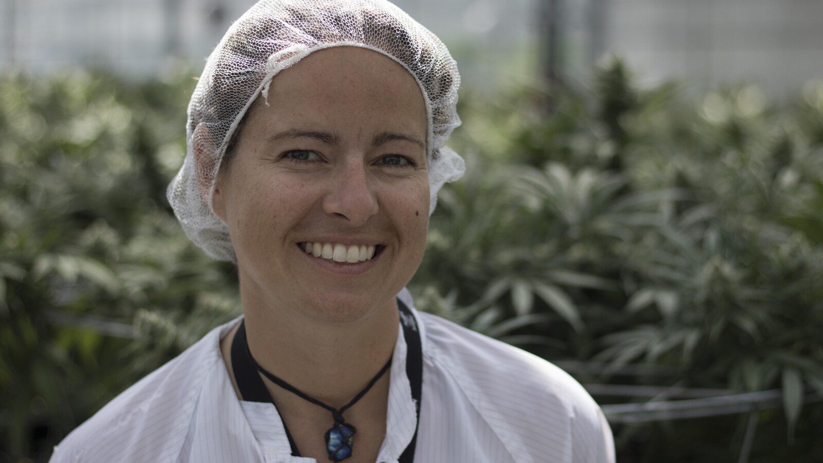 Une jeune femme portant un filet sur les cheveux et un sarrau blanc sourit à la caméra devant des plants de cannabis.