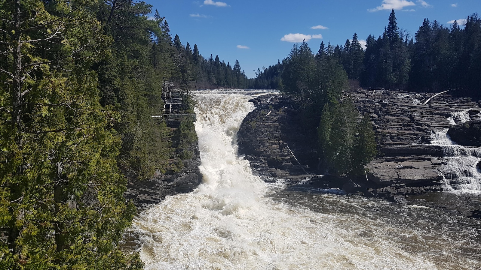 Une immense chute d'eau se déverse dans un bassin naturel. À proximité, on aperçoit un petit escalier de bois pour les visiteurs.