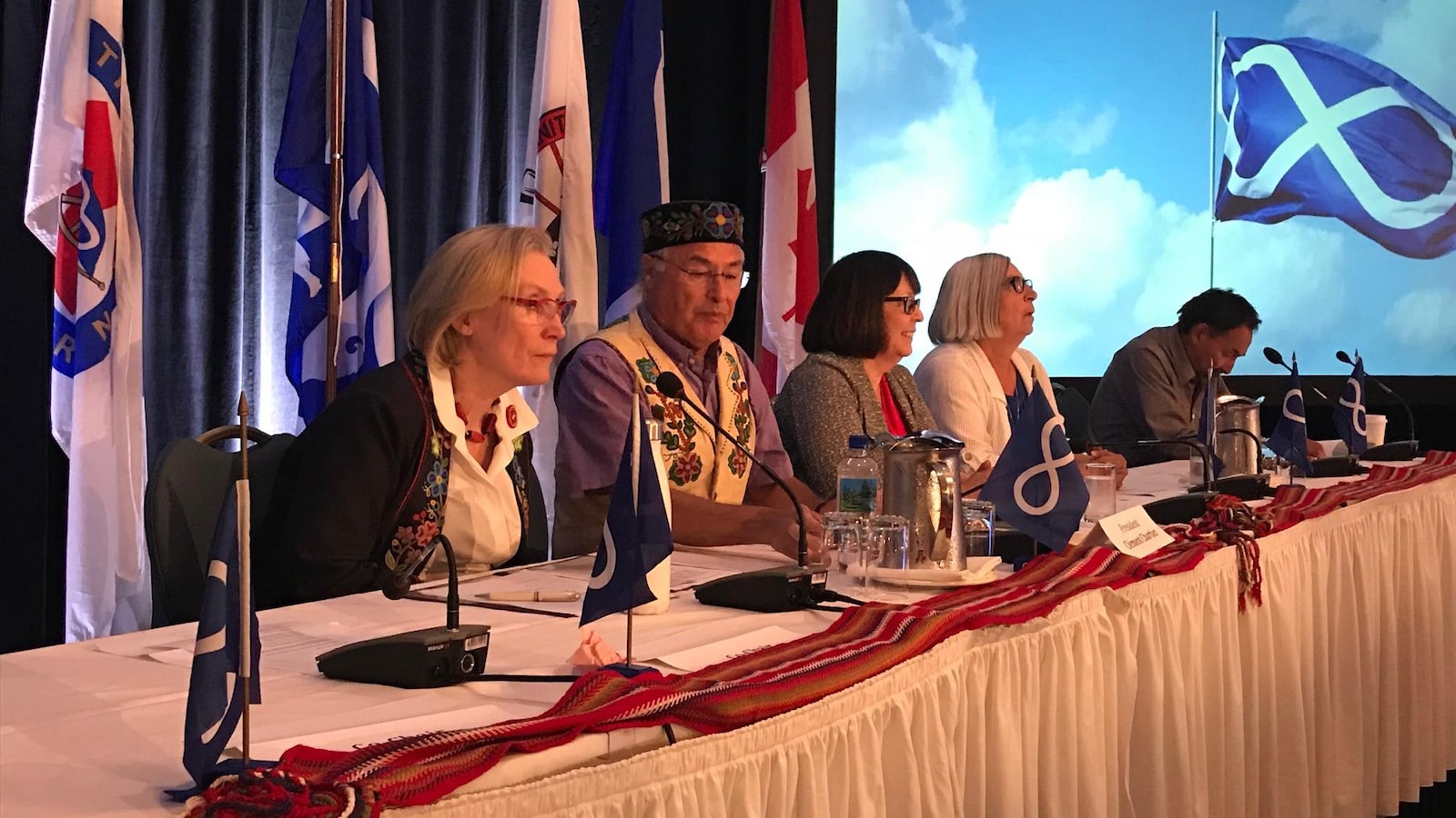 Carolyn Bennett, Clément Chartier et des leaders de la Nation Métisse sont attablés avec des drapeaux métis autour.