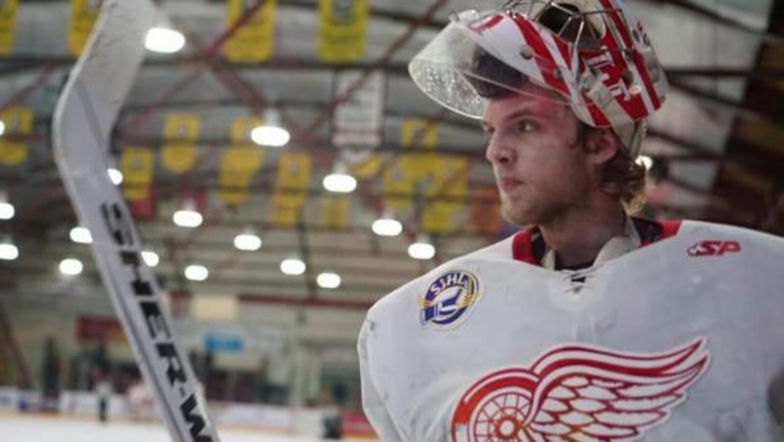 Le joueur en costume, le masque levé, se trouve sur le bord d'une patinoire, son bâton de hockey est aussi levé.