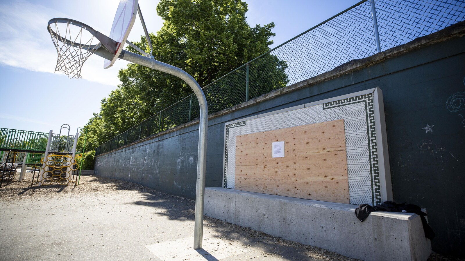Un panneau en bois est apposé sur une plaque en mosaïque, derrière un panier de basketball dans une cour d'école.