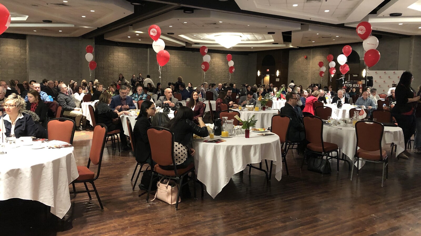 Des personnes sont attablées à des tables rondes dans une salle de conférence décorée avec des ballons rouges et blancs.