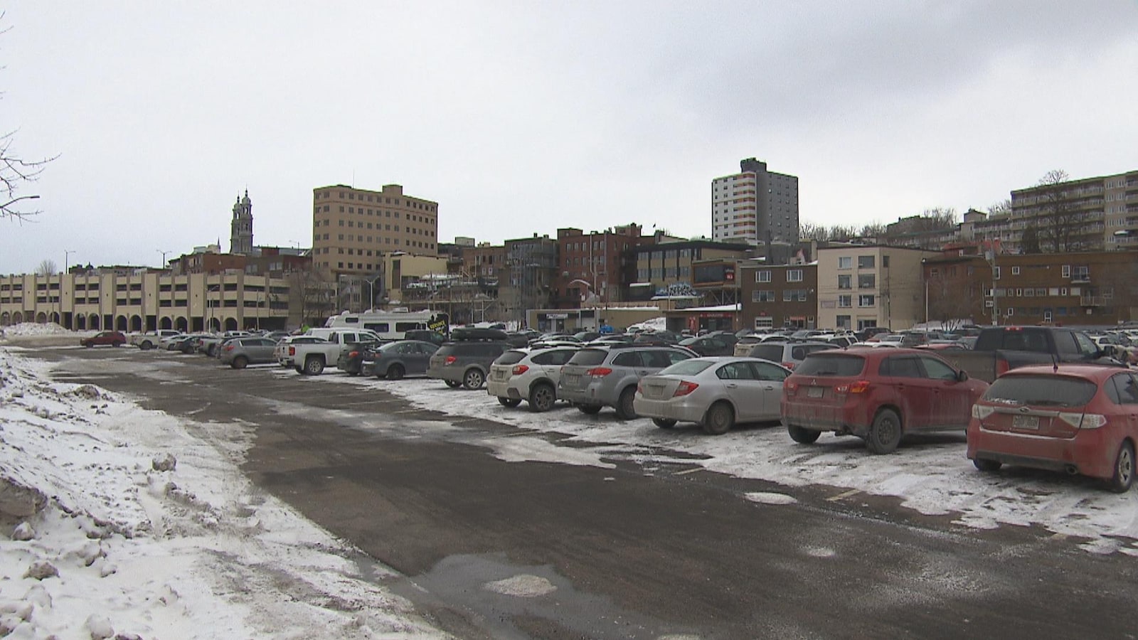Un stationnement avec des véhicules. Derrière, on peut voir le centre-ville de Chicoutimi.
