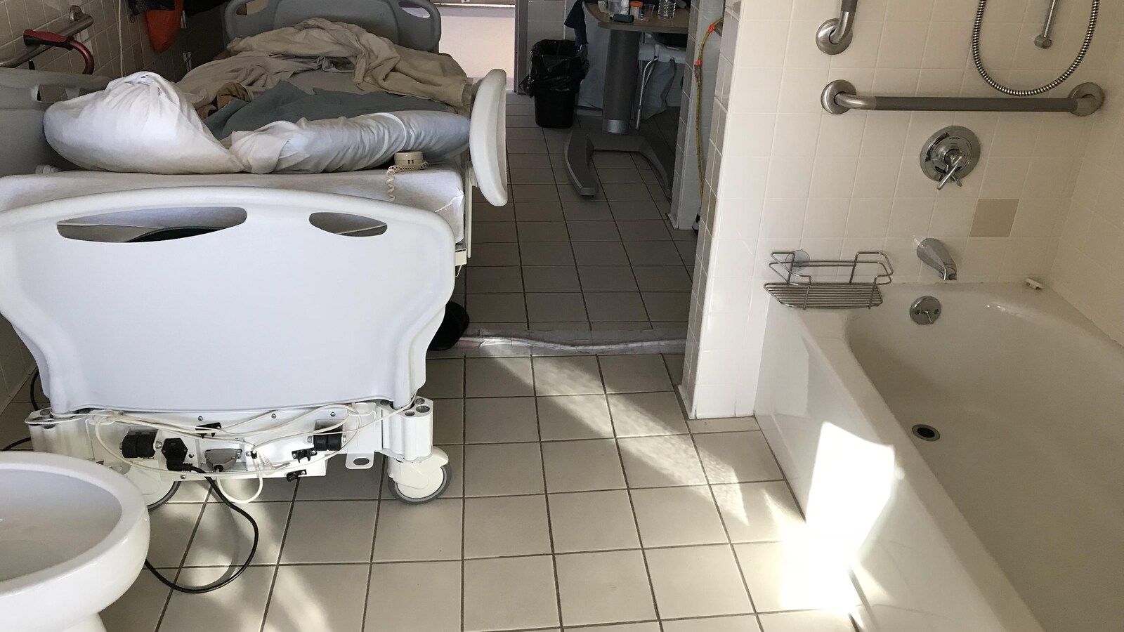 Une salle de bain transformée en chambre d'hôpital. On voit un lit d'hôpital et, au pied du lit, une toilette, et un bain à droite.