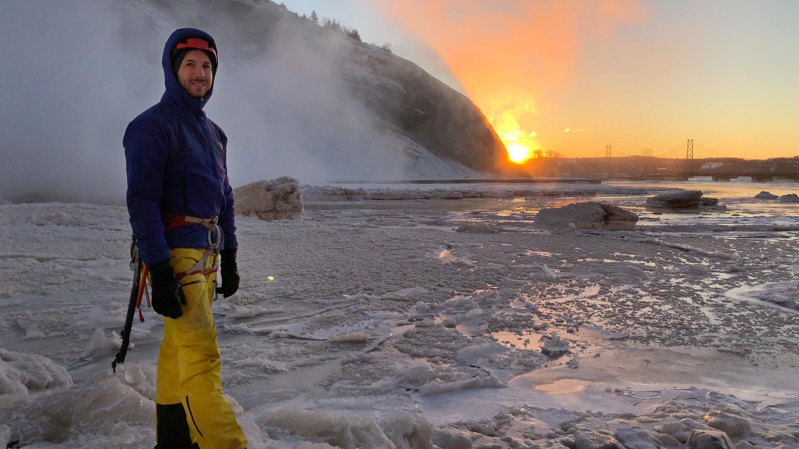 Charles Roberge en équipement d'escalade sur la glace, en bordure des chutes Montmorency et du fleuve, au lever de soleil