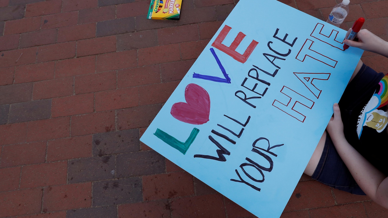 Une personne assise par terre dessine sur un carton sur lequel est écrit : « Love will replace your hate ».