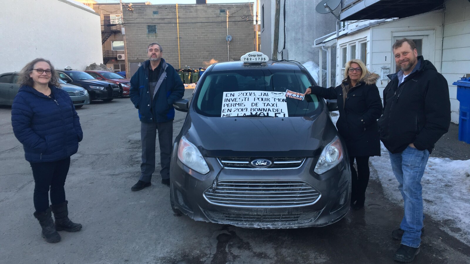 Quatre chauffeurs de taxi se tiennent près d'un taxi. Sur une pancarte il est inscrit « En 2008, j'ai investi pour mon permis de taxi. En 2019 Bonnadel me l'a volé. À vendre zéro dollar ».