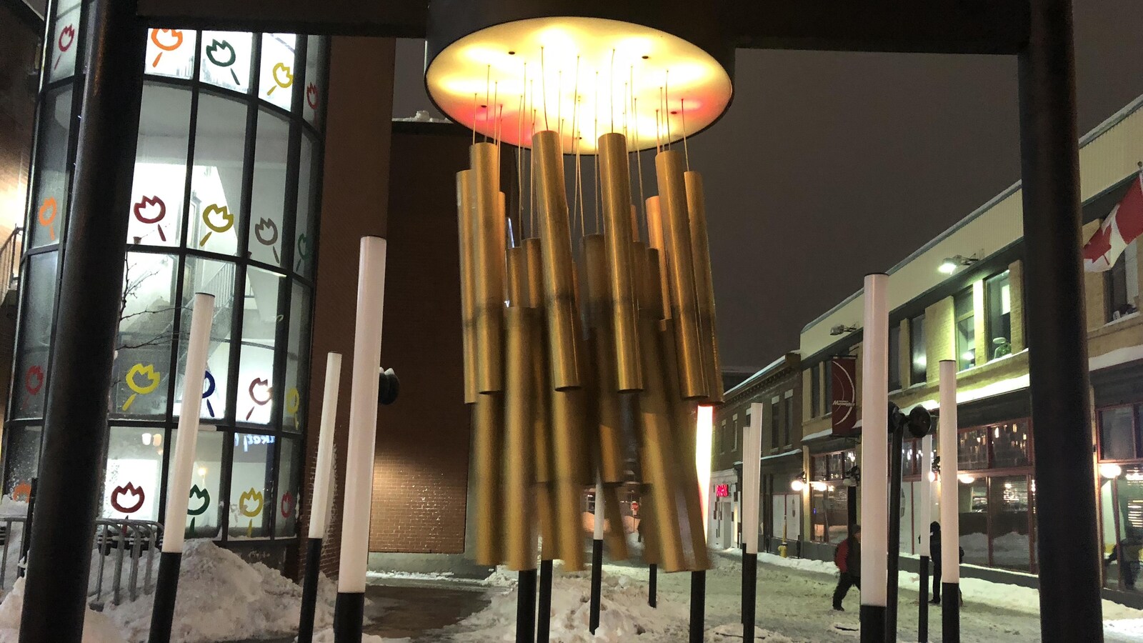 Un carillon est installé entre des colonnes de lumière dans un espace public extérieur enneigé.