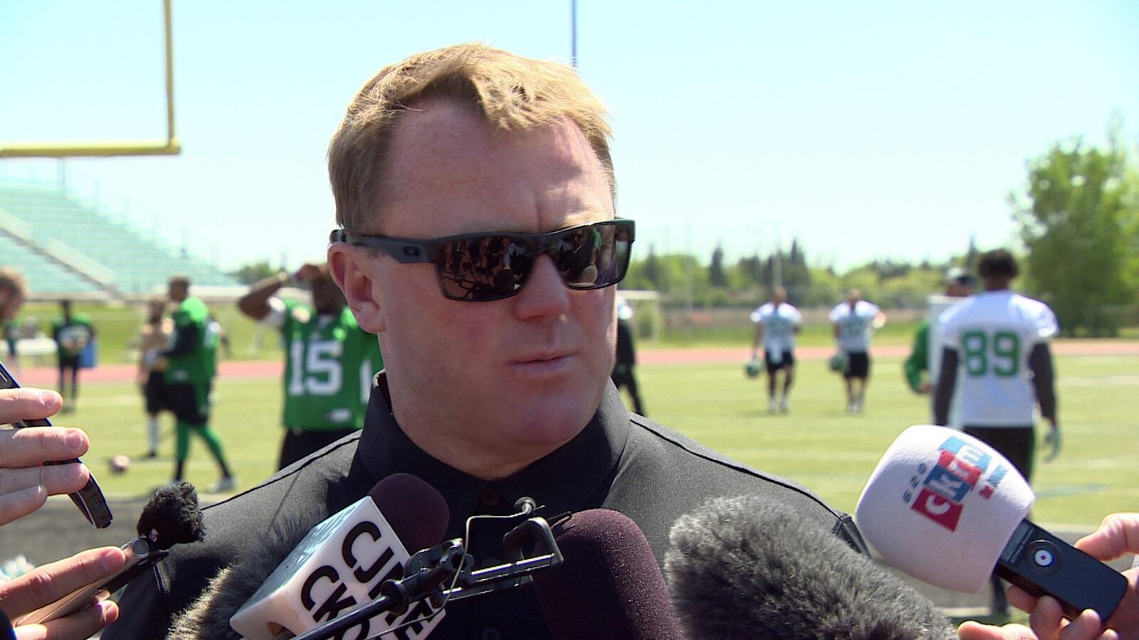 Chris Jones donne une entrevue sur un terrain de football avec les joueurs des Roughriders qui s'entraînent derrière lui.