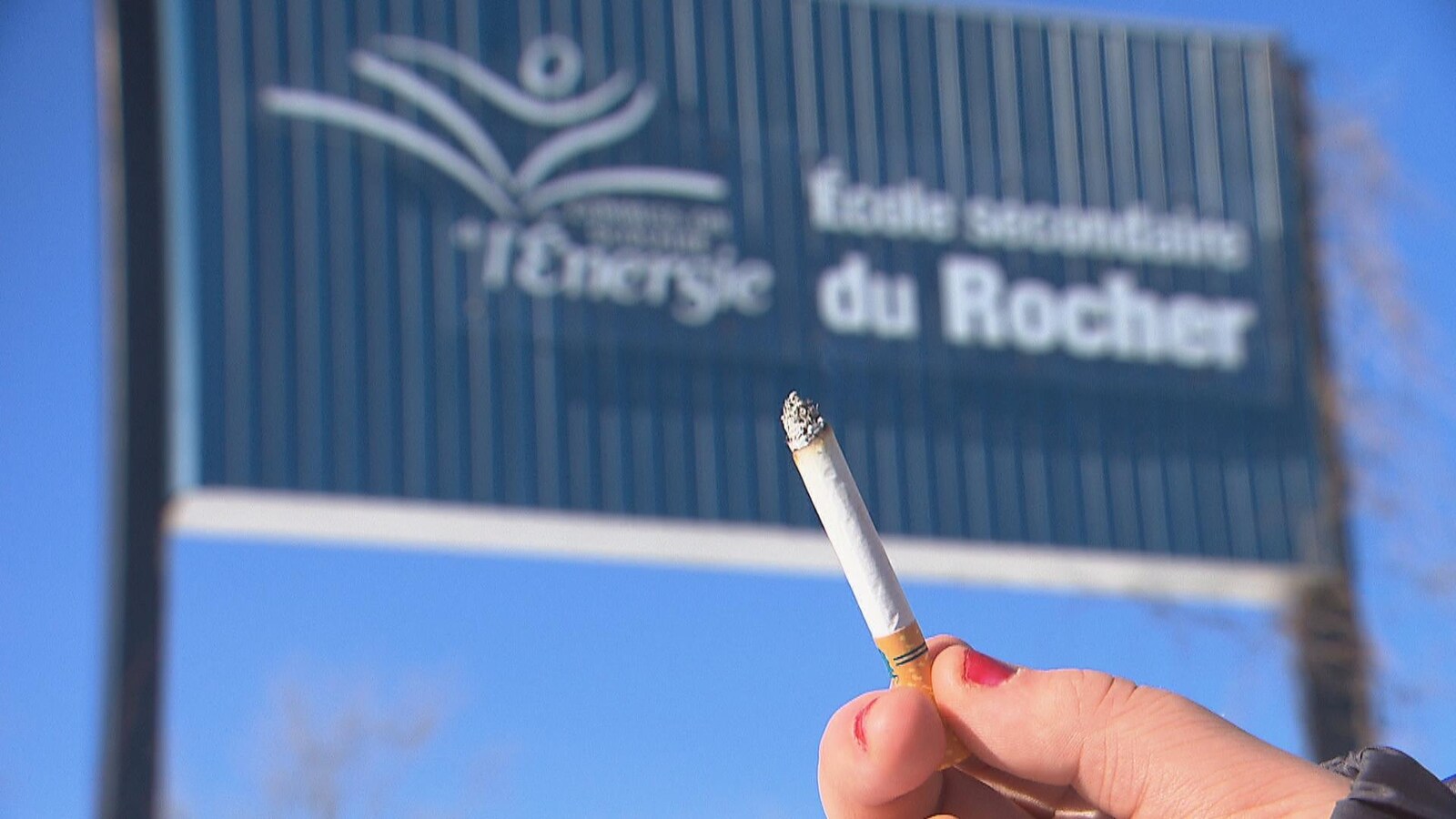 Une femme tient une cigarette devant une affiche d'école.