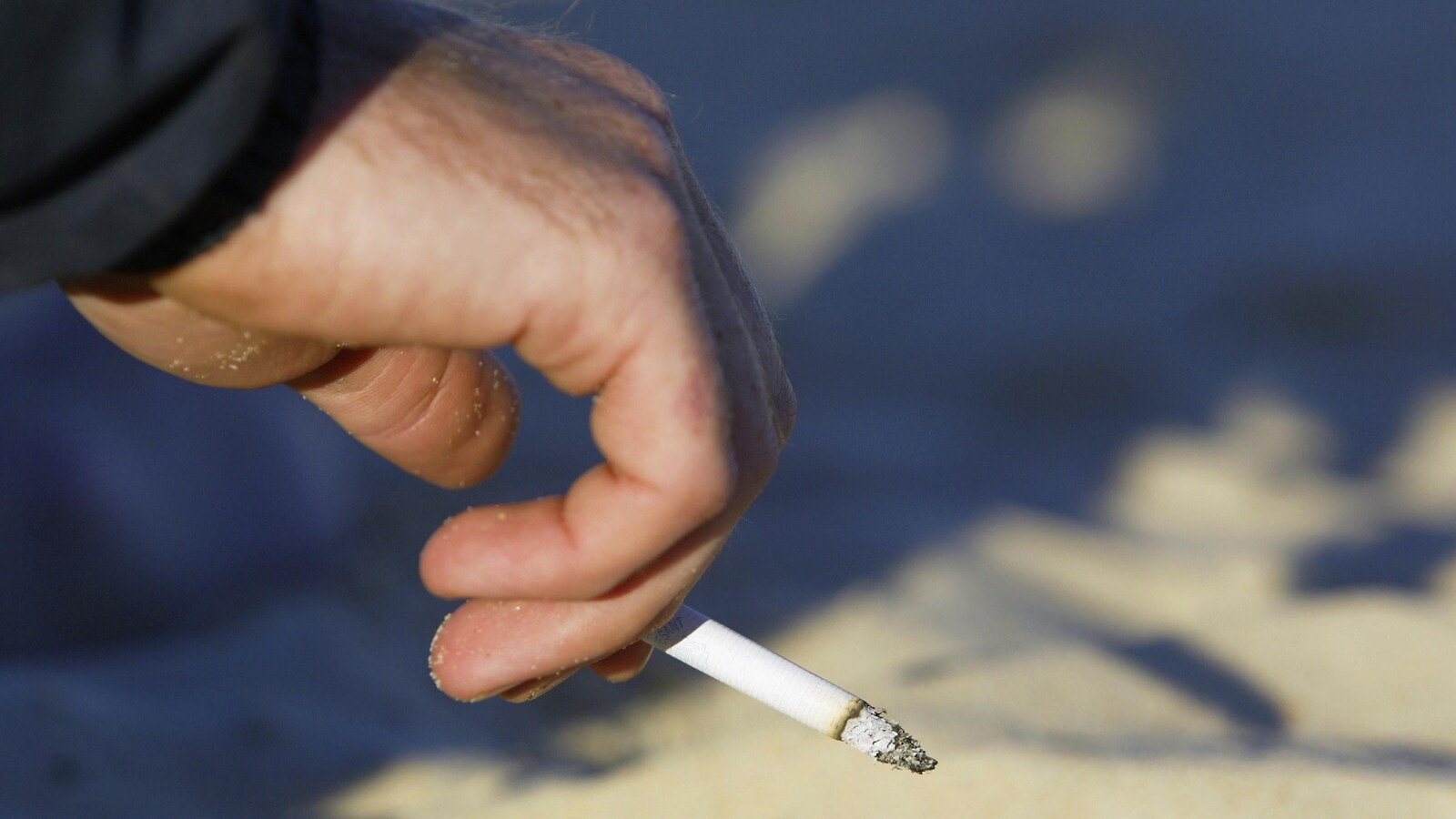 Une personne fume sur la plage. On voit sa main et du sable.