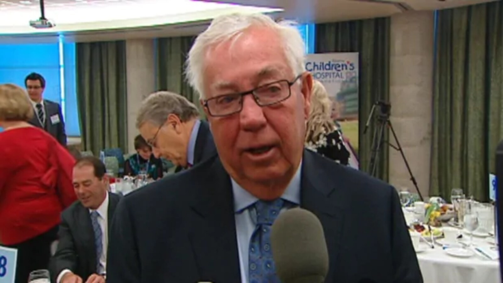 Un homme âgé parle devant un microphone avec, en arrière-plan, des gens attablés.