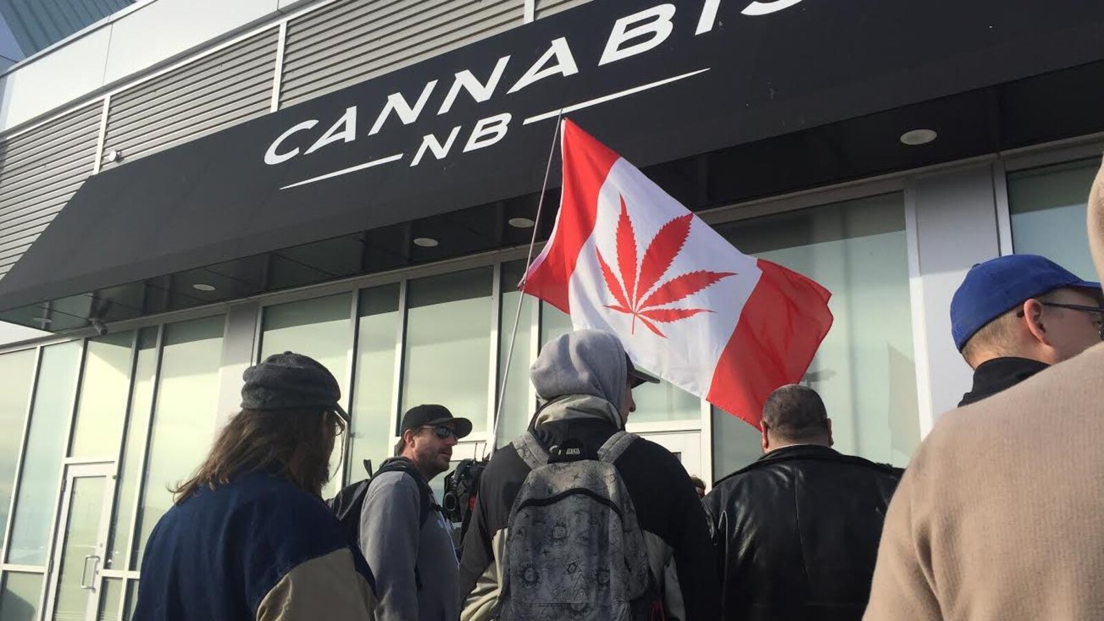 Des clients devant une succursale de Cannabis NB, avec un drapeau canadien où la feuille d'un plant de marijuana remplace la feuille d'érable.