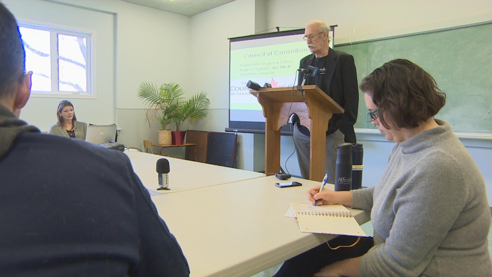 Un homme devant un lutrin parle à des gens qui prennent des notes.