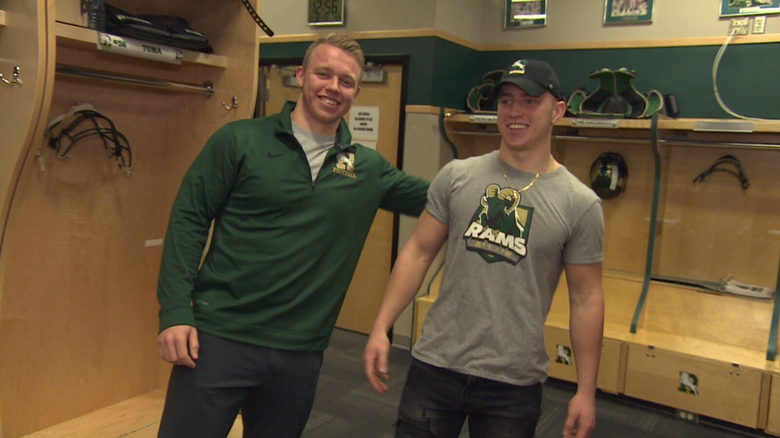 Deux jeunes hommes, le sourire aux lèvres, dans un vestiaire de football avec des vêtements aux couleurs des Rams de l'Université de Regina.