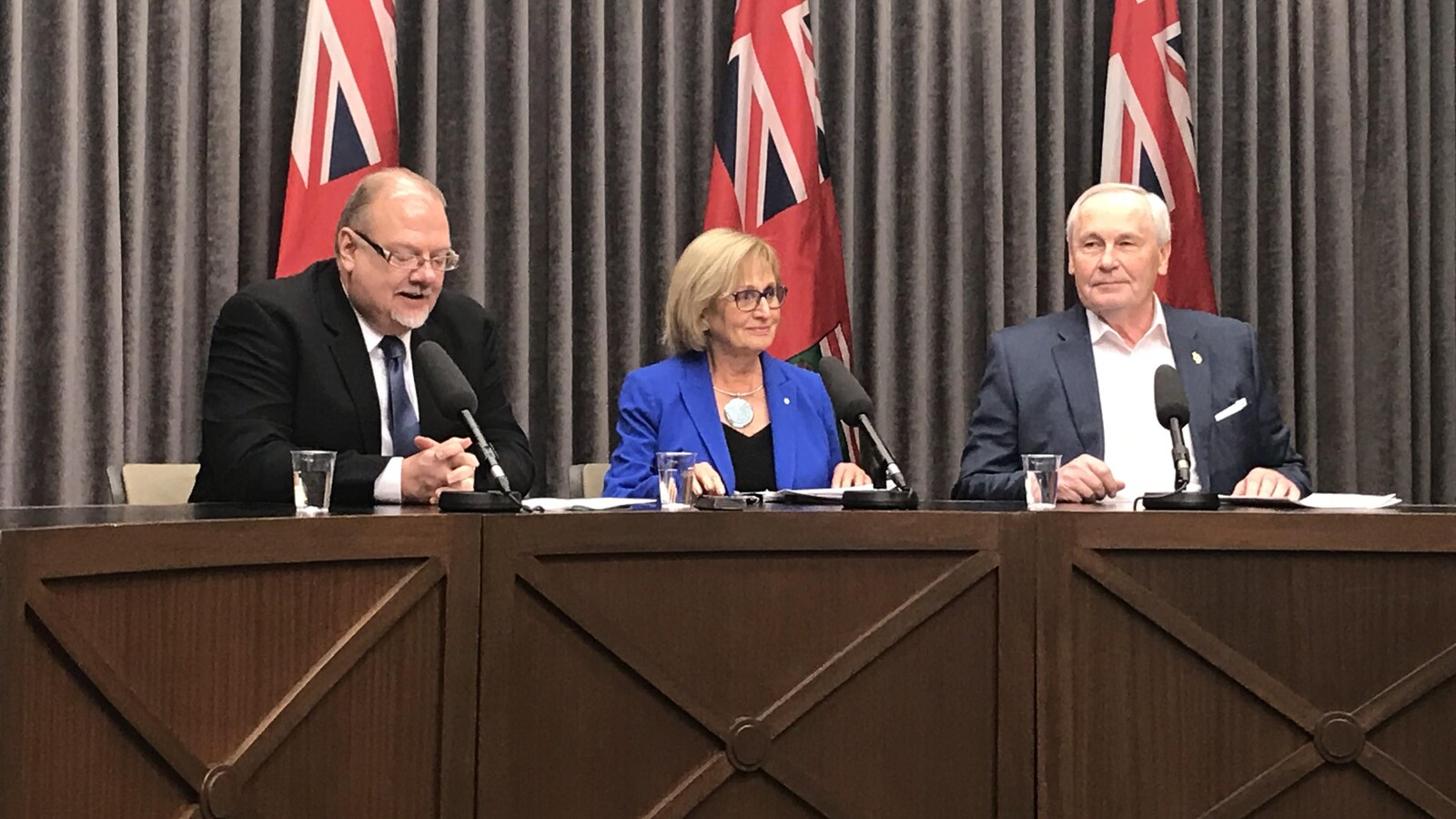 Trois personnes sont assises à une table. L'homme de gauche, le ministre, parle à un micro, pendant que la femme et l'homme situés à sa gauche regardent devant eux et sourient.