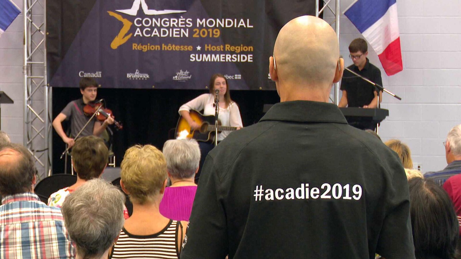 Musiciens sur scène et personne de dos portant un chandail avec le mot-clic #acadie2019.