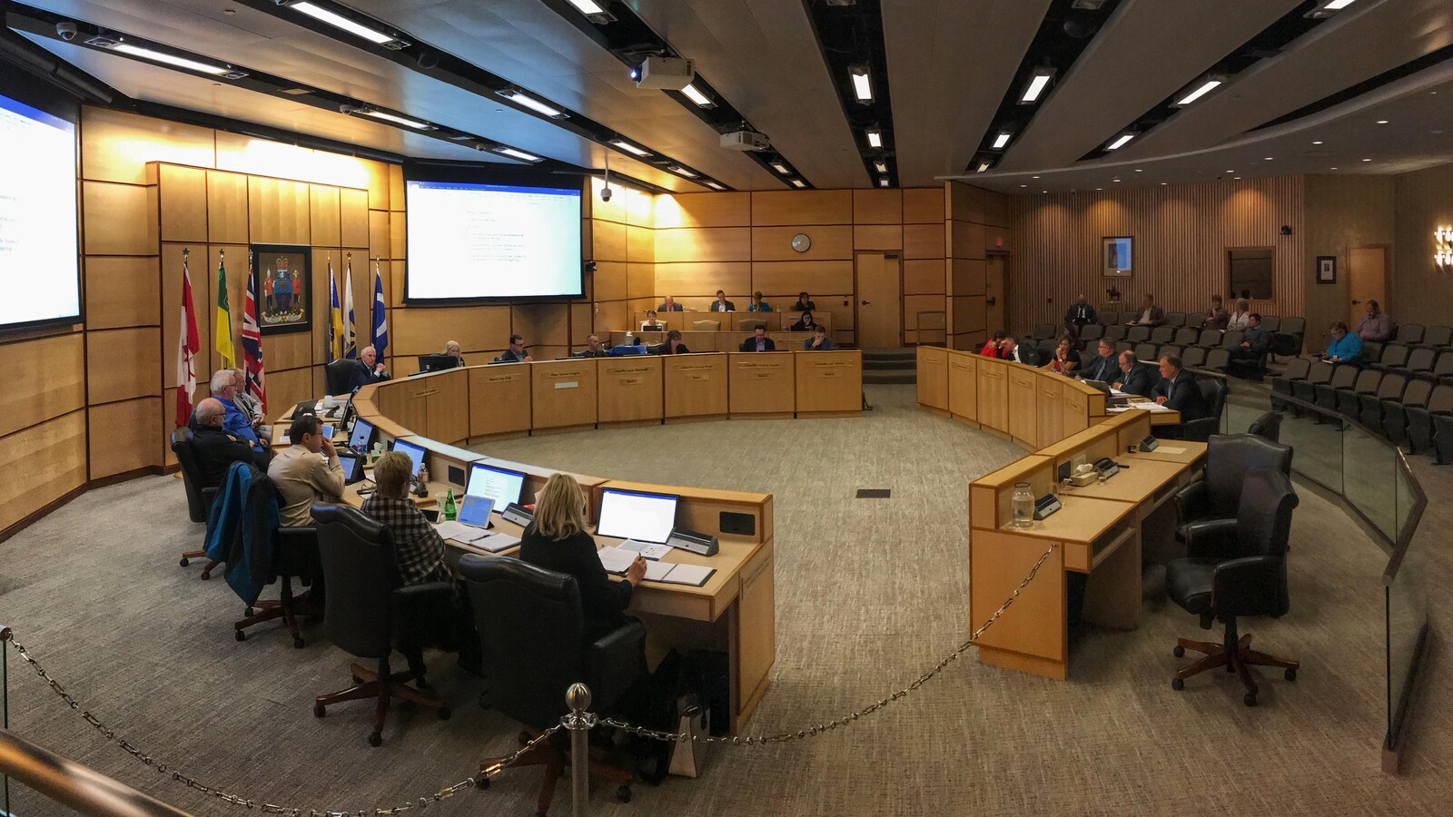 L'intérieur de la salle de rencontre du conseil municipal de la Ville de Regina. Il y a des bureaux installés en demi-cercle avec les conseillers qui sont assis devant un écran.